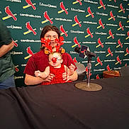 Ayaru is registered to the contest to win money with this photo: baby, child, reindeer_costume, red_nose, antlers, microphone, table, person, red_dress, background, logo, cardinal, christmas, holiday, indoor, event, holding, smile, face, seated