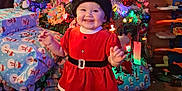 Ayaru joined the competition — help win amazing prizes! baby, christmas_tree, presents, holiday, festive, red_dress, smiling, indoors, decorations, ornaments, santa_hat, child, happy, lights, gift_wrap, floor, carpet, toy, furniture, celebration
