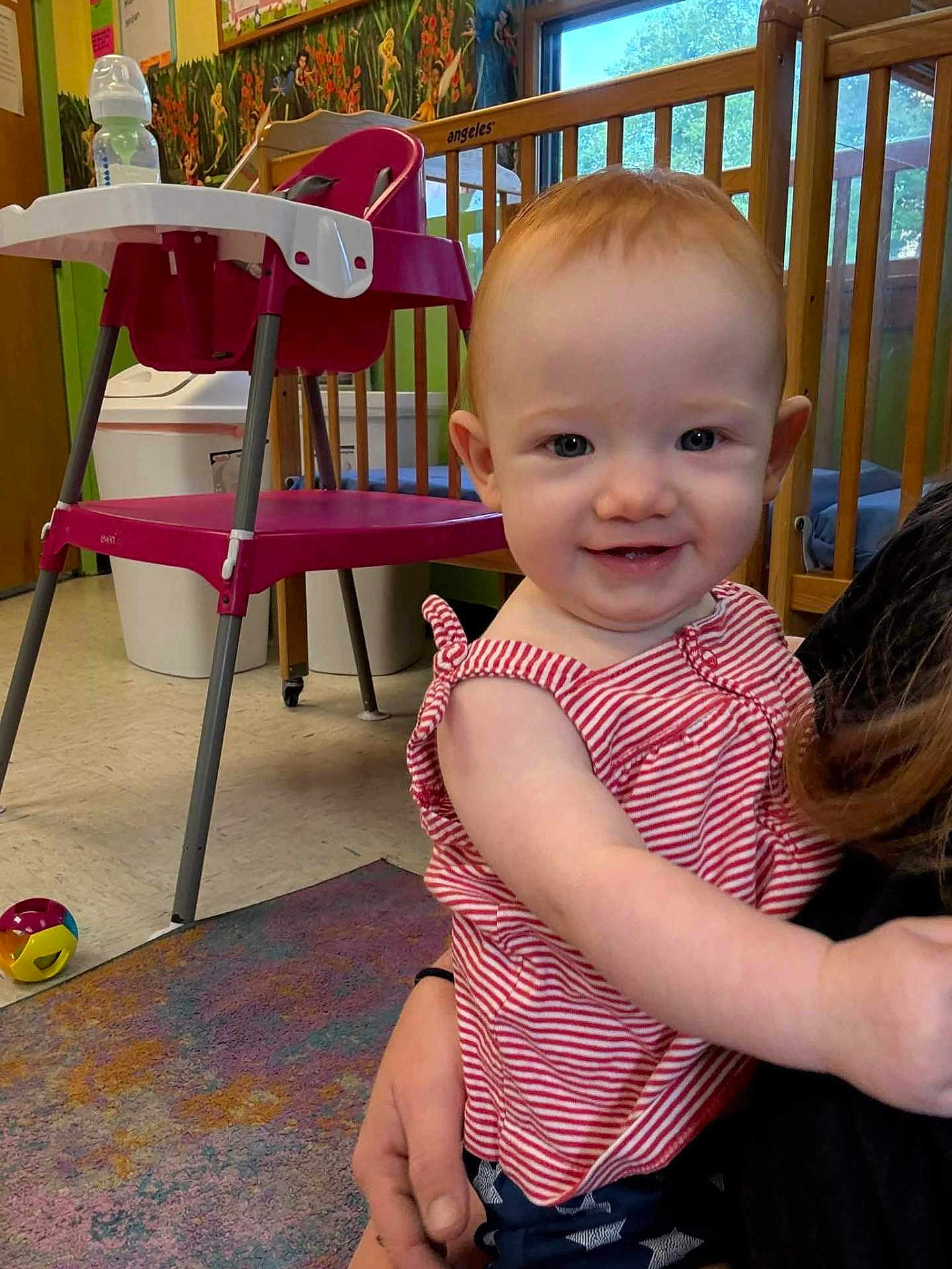 Sophia is registered to the contest to win money with this photo: toddler, child, smiling, red_hair, striped_clothing, indoor, high_chair, ball, carpet, daycare, playroom, person, baby, holding, furniture, window, toy, colorful, happy, face