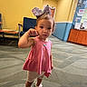 Savannah is registered to the contest to win money with this photo: toddler, child, girl, bow, pink_top, shorts, sandals, indoor, carpet, waiting_room, chair, wall, poster, door, curious, standing, scratching, floor, person, young_child