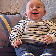 Pierre a rejoint le concours — aidez-le/la à gagner de superbes lots ! baby, smiling, child, striped_shirt, socks, couch, pillow, indoor, happy, seated, cozy, face, hands, feet, clothing, person, portrait, home, baby_toys, pacifier