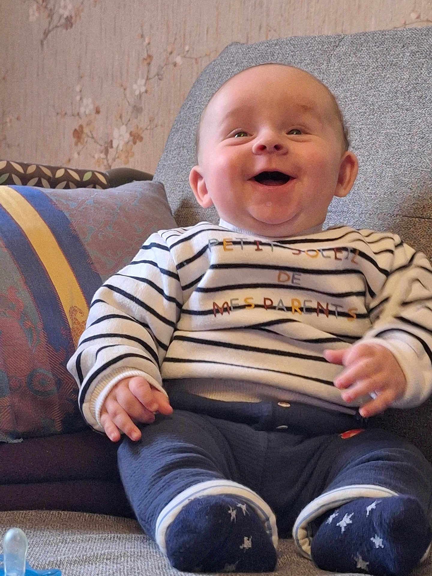 Pierre a rejoint le concours — aidez-le/la à gagner de superbes lots ! baby, smiling, child, striped_shirt, socks, couch, pillow, indoor, happy, seated, cozy, face, hands, feet, clothing, person, portrait, home, baby_toys, pacifier