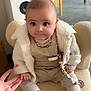 baby, infant, child, plush_chair, white_fleece_jacket, overalls, floral_top, pacifier_clip, beaded_chain, fringe_boots, tights, holding_hand, wide_eyes, chubby_cheeks, cute_expression, indoor, kitchen_background, speaker, floor, soft_lighting