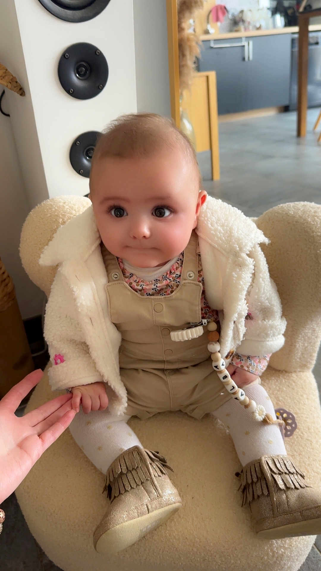 Alba a rejoint le concours — aidez-le/la à gagner de superbes lots ! baby, infant, child, plush_chair, white_fleece_jacket, overalls, floral_top, pacifier_clip, beaded_chain, fringe_boots, tights, holding_hand, wide_eyes, chubby_cheeks, cute_expression, indoor, kitchen_background, speaker, floor, soft_lighting