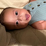 Eliahs is registered to the contest to win money with this photo: baby, tongue, face, eyes, blue_clothing, sleeveless, quilt, closeup, infant, cute, skin, button, lying_down, playful, child, person, indoors, expression, head, baby_garment