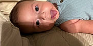 Eliahs is registered to the contest to win money with this photo: baby, tongue, face, eyes, blue_clothing, sleeveless, quilt, closeup, infant, cute, skin, button, lying_down, playful, child, person, indoors, expression, head, baby_garment