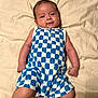 baby, infant, smiling, blue_clothing, white_clothing, checkered_pattern, romper, barefoot, ankle_bracelet, lying_down, bed_sheet, beige_background, cute, child, person, young, skin, face, happy, indoors