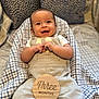 Eliahs is registered to the contest to win money with this photo: baby, infant, smiling, overalls, blanket, checkered, cute, happy, clothing, face, child, person, indoor, portrait, sitting, hands, head, cushion, fabric, three_months