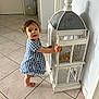 child, toddler, blue_dress, barefoot, tile_floor, birdcage, decor, indoor, standing, curious, short_hair, home, person, young_child, looking_up, holding, white_birdcage, furniture, light_wall, door