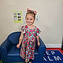 blue_couch, bow, carpet, child, classroom, colorful_clothing, cute, dress, educational_posters, floor, girl, happy, interior, person, smiling, sneakers, standing, tattoos, wall, young