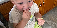 Waylon is registered to the contest to win money with this photo: toddler, child, blond_hair, eating, snack, shorts, dinosaur_print, shirt, sitting, chair, kitchen, cabinets, floor, tile_floor, footwear, sandals, light, indoor, curious, person