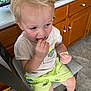 toddler, child, blond_hair, eating, snack, shorts, dinosaur_print, shirt, sitting, chair, kitchen, cabinets, floor, tile_floor, footwear, sandals, light, indoor, curious, person