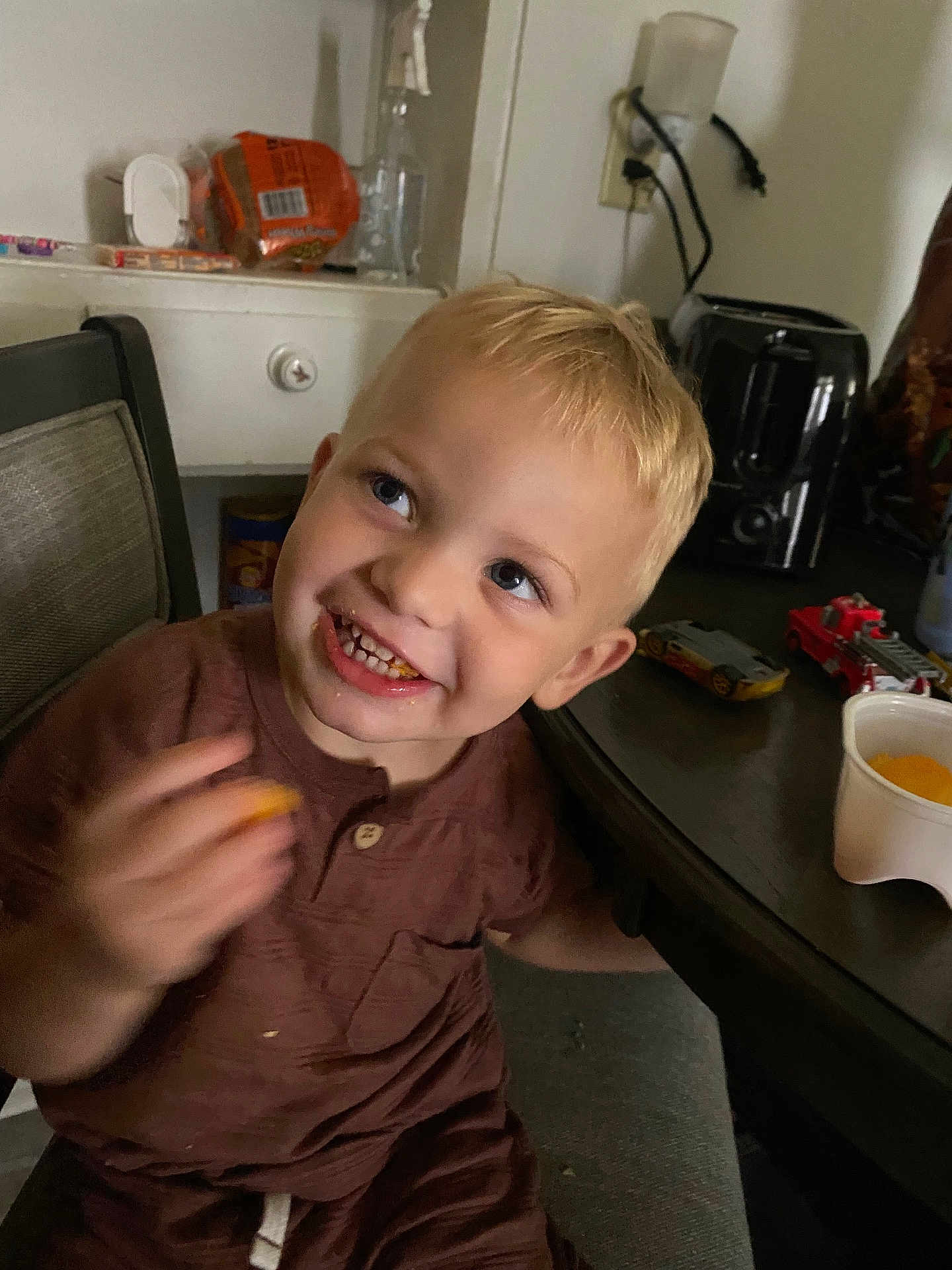 Waylon joined the competition — help win amazing prizes! child, toddler, blond_hair, smiling, snacking, brown_clothing, table, toy_cars, cup, kitchen, chair, face, happy, indoor, food, cheese_puffs, person, cute, messy, playful