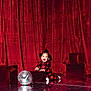 baby, bodypart, clothing, couch, dress, electricaldevice, face, finger, furniture, hand, head, lighting, microphone, performer, person, photography, portrait, soloperformance, stage, velvet