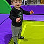 child, toddler, smiling, standing, trampoline, playground, indoor, colorful, fun, happy, black_shirt, gray_pants, socks, netting, play_area, young_child, casual_clothing, short_hair, active, recreation