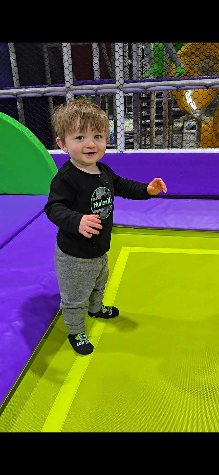 Maxton joined the competition — help win amazing prizes! child, toddler, smiling, standing, trampoline, playground, indoor, colorful, fun, happy, black_shirt, gray_pants, socks, netting, play_area, young_child, casual_clothing, short_hair, active, recreation