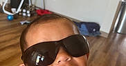 Kamaka is registered to the contest to win money with this photo: child, toddler, smiling, sunglasses, portrait, close_up, indoor, home, living_room, kitchen, high_chair, wood_floor, toys, furniture, selfie, face, head, shoulders, playful, casual