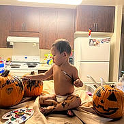 Kamaka is registered to the contest to win money with this photo: child, toddler, baby, diaper, sitting, painting, paintbrush, pumpkin, jack_o_lantern, halloween, kitchen, indoor, refrigerator, stove, sink, countertop, art_supplies, palette, creative, portrait