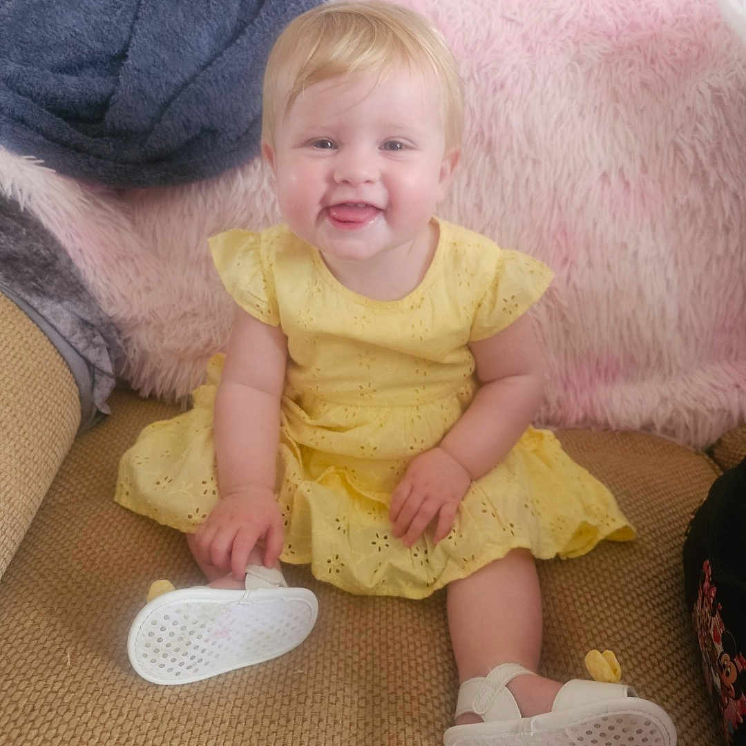 Evie-Jo joined the competition — help win amazing prizes! baby, blanket, blonde_hair, casual, child, couch, cozy, cute, furniture, happy, indoor, infant, person, playful, portrait, sitting, smiling, soft_texture, white_sandals, yellow_dress