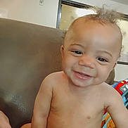 Messiah is registered to the contest to win money with this photo: baby, smiling, child, curly_hair, shirtless, couch, indoor, diaper, happy, infant, person, face, skin, expression, childhood, cute, portrait, home, furniture, blanket