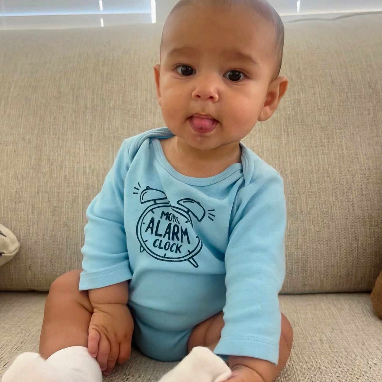 Zylen is registered to the contest to win money with this photo: baby, infant, child, onesie, blue_clothing, socks, couch, sofa, indoor, cute, tongue_out, sitting, person, young_child, smiling, home, furniture, window_blinds, skin, head