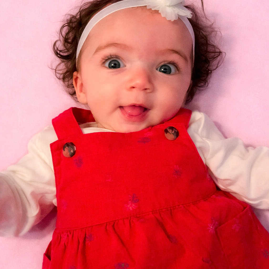 Lillyonna is registered to the contest to win money with this photo: baby, infant, child, curly_hair, headband, flower_headband, red_dress, white_shirt, smiling, tongue_out, lying_down, pink_background, cute, portrait, happy, one_person, indoors, closeup, baby_fashion, adorable