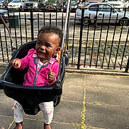 Kaylani is registered to the contest to win money with this photo: child, toddler, swing, playground, park, smiling, happy, black_hair, pink_jacket, leggings, barefoot, outdoor, daylight, fence, cars, sidewalk, metal, joy, fun, person