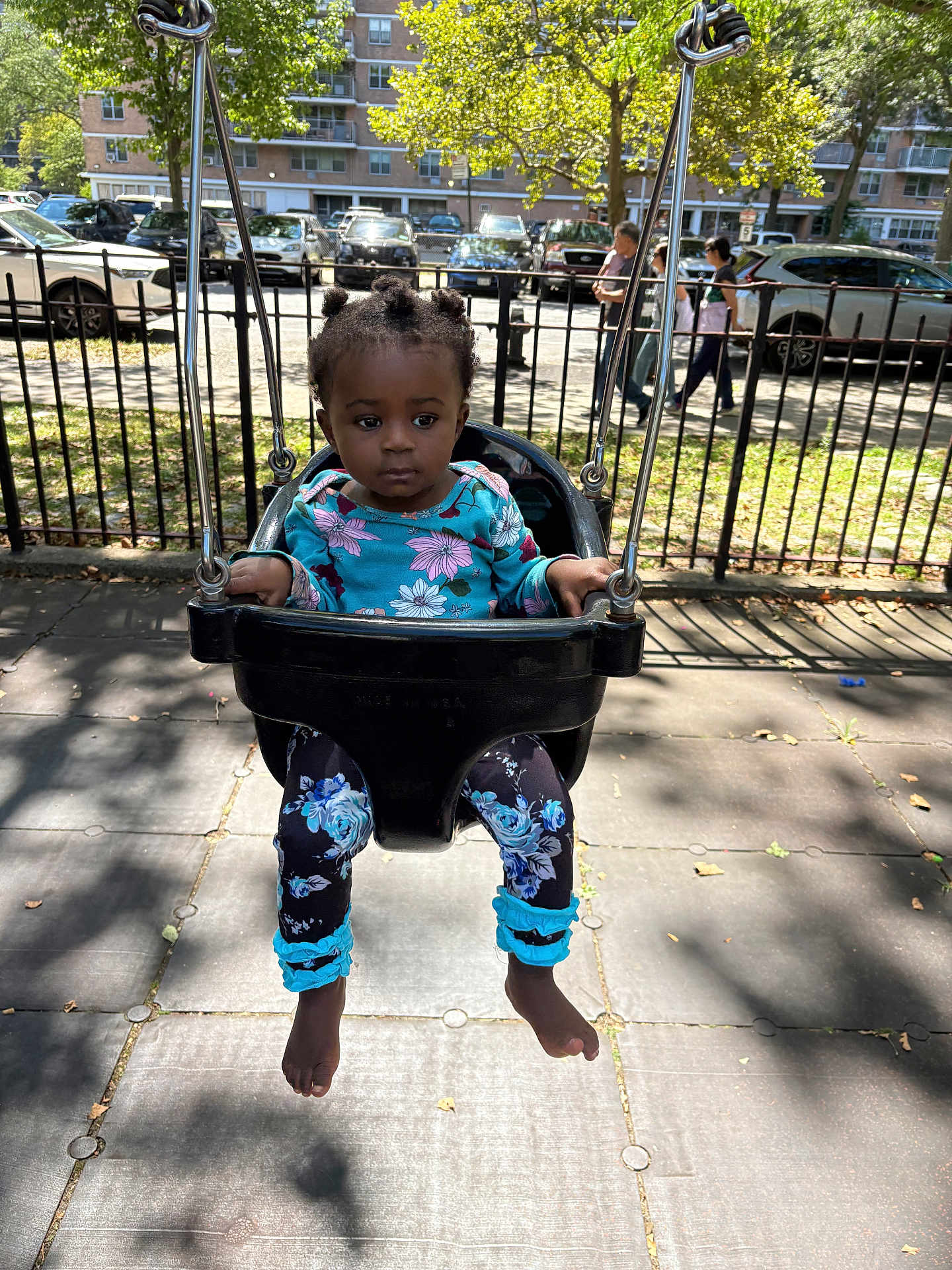 Kaylani joined the competition — help win amazing prizes! toddler, child, swing, playground, outdoor, park, black_swing, floral_clothing, barefoot, person, urban, daylight, trees, sidewalk, fence, people, car, shadow, expression, young_child