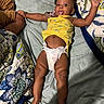 adult_hand, baby, bed, child, comfort, cute, diaper, fabric, floral_pattern, hand, indoors, infant, lying_down, person, playful, quilt, skin, sleepwear, tongue_out, yellow_top