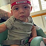 adult_arm, baby, car_seat, child, close_up, gray_clothing, harness, headband, indoors, infant, looking_at_camera, portrait, safe, serious_expression, short_sleeves, sitting, sunlight, tattoo, vehicle_interior, window