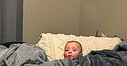 Eli is registered to the contest to win money with this photo: baby, infant, child, face, portrait, blanket, bed, pillow, bedding, gray_blanket, cozy, indoors, home, sitting, peeking, looking_at_camera, neutral_expression, textile, soft_furnishings, photography