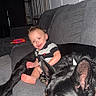 animal, baby, bare_feet, black_dog, child, companion, couch, dog, happy, home, indoor, large_dog, living_room, pet, portrait, sitting, smiling, sofa, toddler, toy