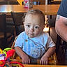 Eli is registered to the contest to win money with this photo: baby, child, toddler, sitting, high_chair, portrait, wide_eyed, serious_expression, light_hair, overalls, white_shirt, toy, colorful_toy, restaurant, table, chair, indoor, sunlight, adult, food_packaging