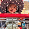 baby, child, eyes, face, family_outing, green_clothing, happy, indoor, infant, open_mouth, portrait, red_cart, seated, shopping, shopping_cart, smiling, star_pattern, store, sun_hat, supermarket