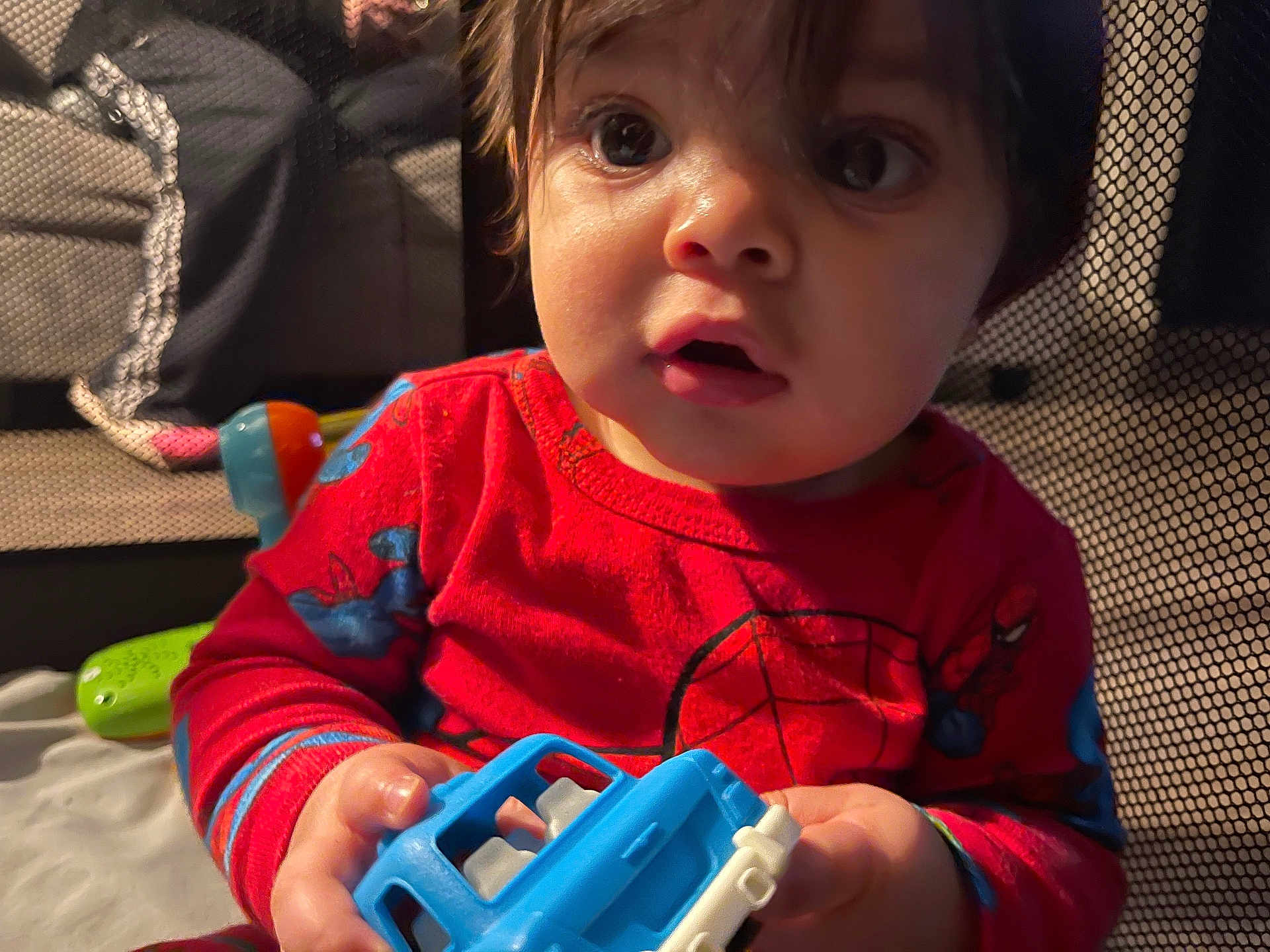 Cartier is registered to the contest to win money with this photo: baby, blue_toy, candid, child, closeup, curious, expression, face, hands, indoor, mesh, pajamas, playing, playpen, red_clothing, soft_lighting, spiderman, toddler, toy, wide_eyes
