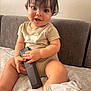 Cartier is registered to the contest to win money with this photo: baby, child, remote_control, couch, blanket, indoor, happy, smiling, person, toddler, clothing, sitting, furniture, cute, skin, hand, face, hair, room, relaxed