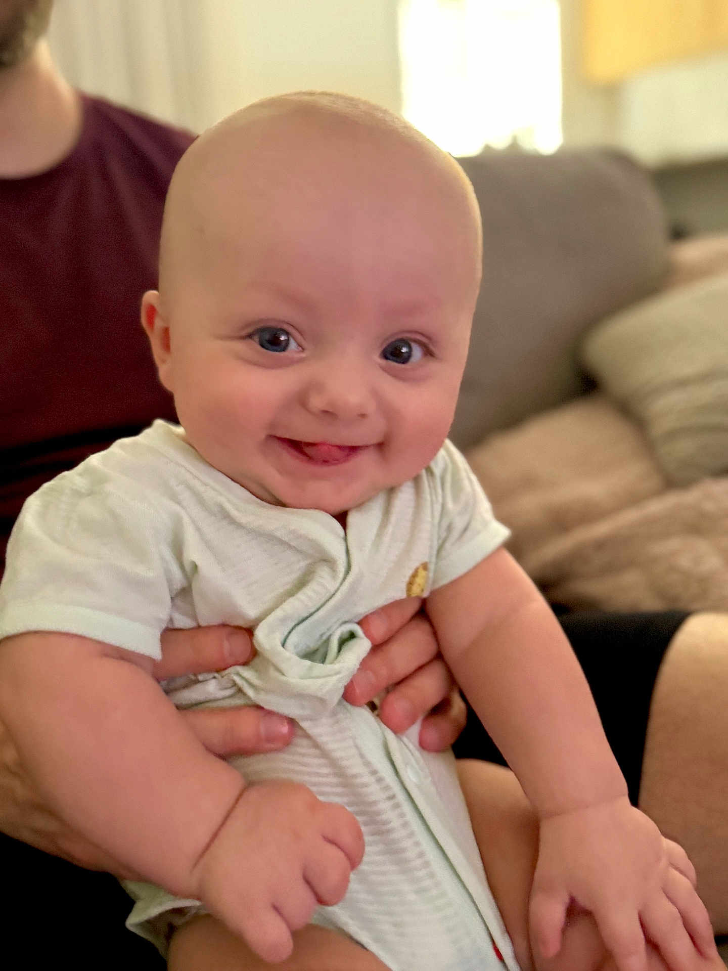 Nathaniel is registered to the contest to win money with this photo: baby, smiling, tongue_out, infant, person, hand, clothing, seated, indoor, couch, adult_hand, cute, happy, portrait, face, skin, young_child, cozy, home, bright_eyes