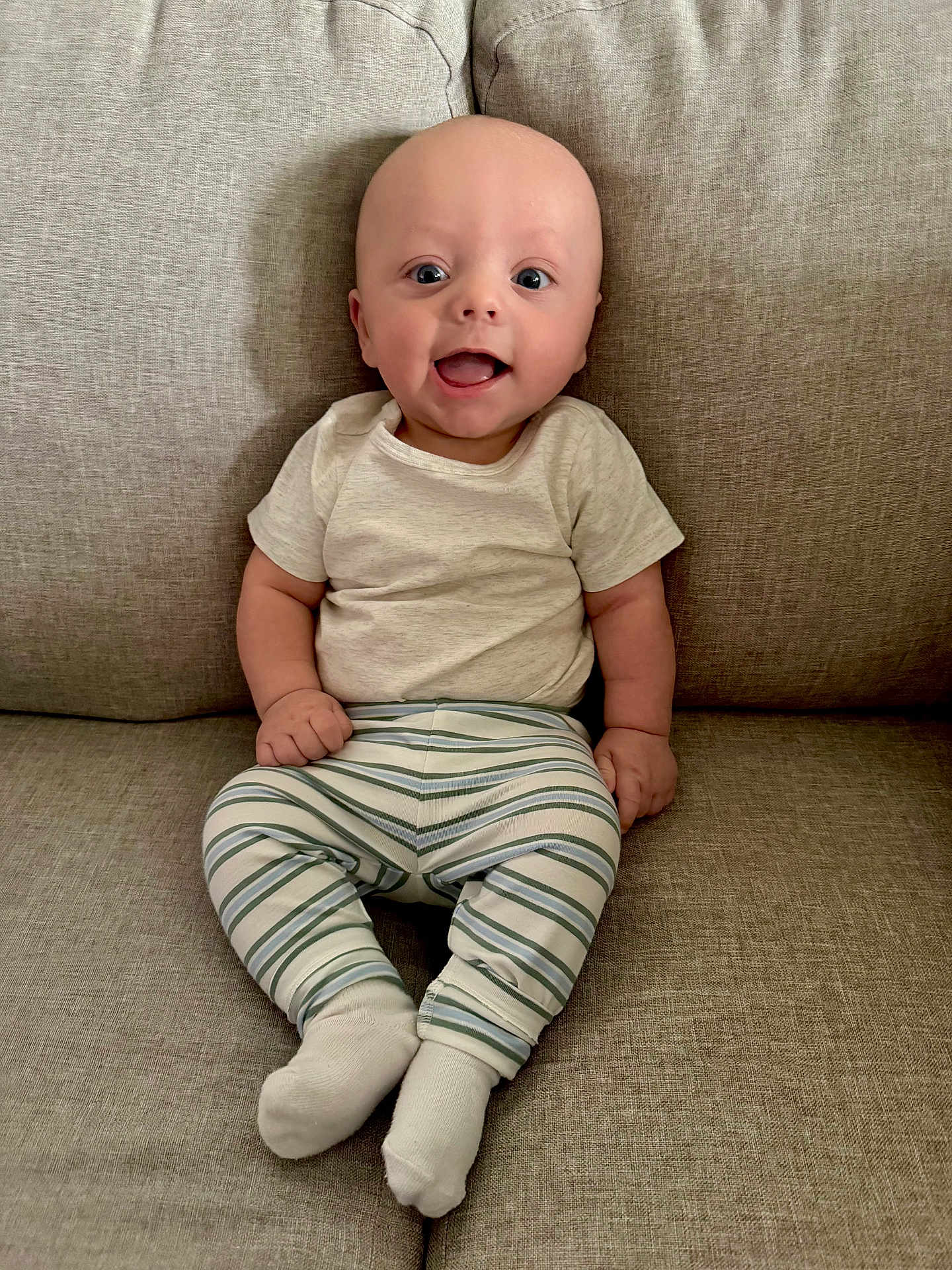 Nathaniel is registered to the contest to win money with this photo: baby, infant, smiling, sitting, couch, striped_pants, white_shirt, socks, happy, indoors, child, face, human, cute, young, portrait, furniture, comfort, casual, expression