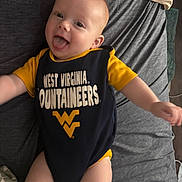 Daniel joined the competition — help win amazing prizes! baby, infant, smiling, tongue_out, clothing, onesie, bed, bedding, pillow, person, child, cute, happy, indoors, laying, yellow, navy_blue, text_on_clothing, face, skin