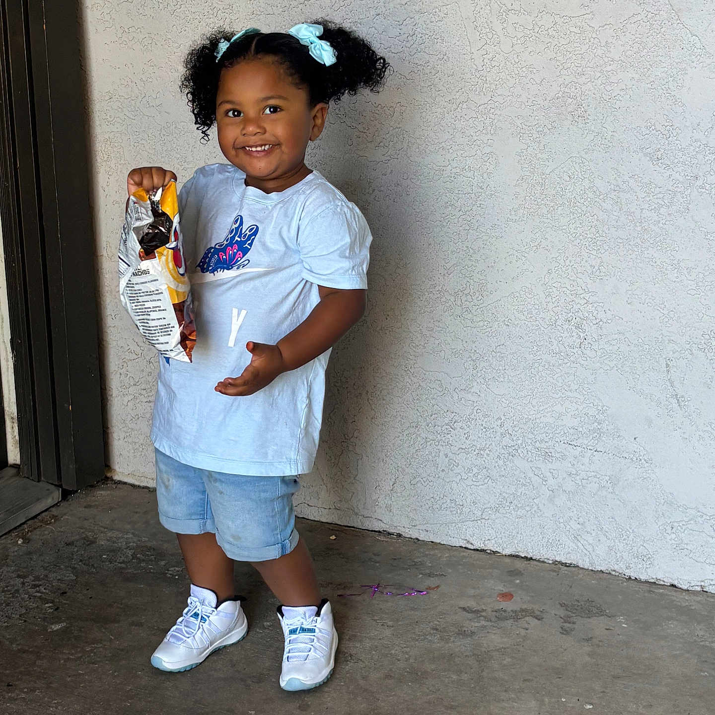 Janiece joined the competition — help win amazing prizes! casual_clothing, child, chips, concrete_floor, curly_hair, cute, denim_shorts, door_frame, hair_bows, happy, indoor, person, portrait, smiling, snack, standing, toddler, wall, white_shoes, white_tshirt