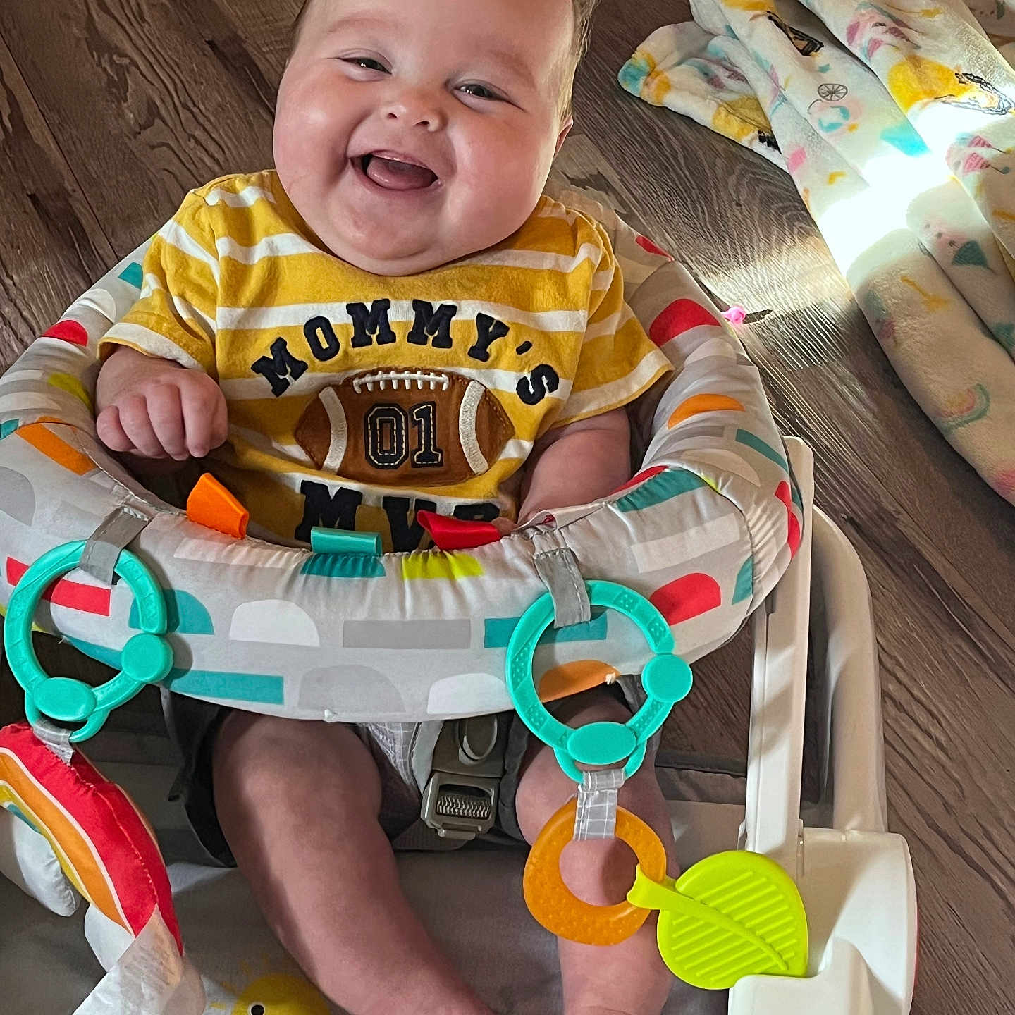 Kamden is registered to the contest to win money with this photo: baby, baby_seat, blanket, child, clothing, colorful, cute, face, feet, floor, happy, indoors, infant, person, play, seat, smiling, teether, toy, wood_floor