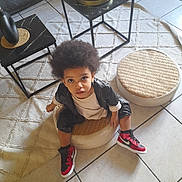 Maēl a rejoint le concours — aidez-le/la à gagner de superbes lots ! afro_hair, black_jacket, casual_clothing, child, coffee_table, curious_expression, home_interior, indoor, looking_up, modern_furniture, plant, red_shoes, round_cushion, rug, sitting, sneakers, tile_floor, toddler, woven_cushion, young_child