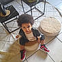 afro_hair, black_jacket, casual_clothing, child, coffee_table, curious_expression, home_interior, indoor, looking_up, modern_furniture, plant, red_shoes, round_cushion, rug, sitting, sneakers, tile_floor, toddler, woven_cushion, young_child