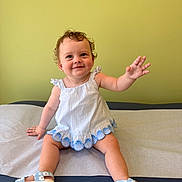 Sophie is registered to the contest to win money with this photo: baby, casual, child, curly_hair, cute, dress, footwear, green_wall, happy, indoor, infant, padded_surface, person, playful, portrait, sandals, sitting, smiling, waving_hand, young_child