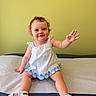 baby, child, smiling, sitting, dress, sandals, curly_hair, waving_hand, indoor, green_wall, padded_surface, cute, happy, portrait, young_child, footwear, infant, person, casual, playful