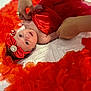 baby, infant, red_bow, headband, red_cloth, flower_petals, white_bedspread, hands, rings, portrait, newborn, cute, lying_down, soft_lighting, closeup, skin, decorative, colorful, celebration, adorable