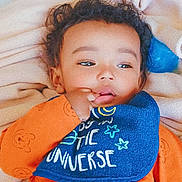 Marcelino is registered to the contest to win money with this photo: baby, child, orange_clothing, bib, blue_bib, blanket, curly_hair, lying_down, indoor, cute, infant, toddler, face, hand, sucking_thumb, soft_texture, sleepwear, portrait, young_child, cozy