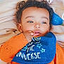 baby, child, orange_clothing, bib, blue_bib, blanket, curly_hair, lying_down, indoor, cute, infant, toddler, face, hand, sucking_thumb, soft_texture, sleepwear, portrait, young_child, cozy