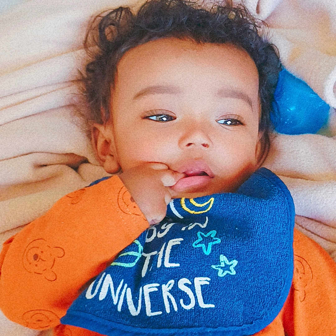 Marcelino is registered to the contest to win money with this photo: baby, bib, blanket, blue_bib, child, cozy, curly_hair, cute, face, hand, indoor, infant, lying_down, orange_clothing, portrait, sleepwear, soft_texture, sucking_thumb, toddler, young_child