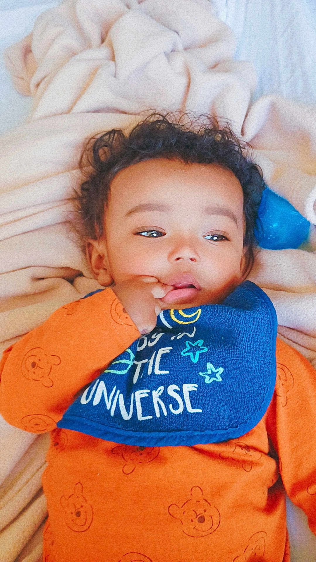 Marcelino is registered to the contest to win money with this photo: baby, child, orange_clothing, bib, blue_bib, blanket, curly_hair, lying_down, indoor, cute, infant, toddler, face, hand, sucking_thumb, soft_texture, sleepwear, portrait, young_child, cozy